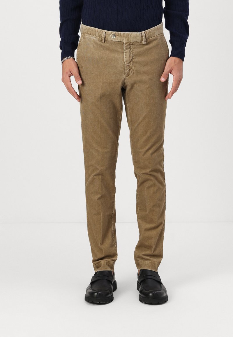 Pantalons en velours côtelé tan avec une coupe droite, dotés d'un bouton et de passants de ceinture. Portés avec des chaussures noires.