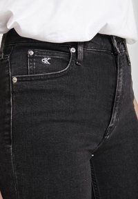 Jeans de mezclilla negra con un diseño ajustado, que presentan un bolsillo frontal con un logo blanco, herrajes plateados y una textura suave.