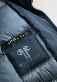 Intérieur d'une veste matelassée bleu marine avec une étiquette noire portant l'inscription "BAKER by TED BAKER" et un graphique pour une "POCHE TECHNOLOGIQUE". Texture lisse.