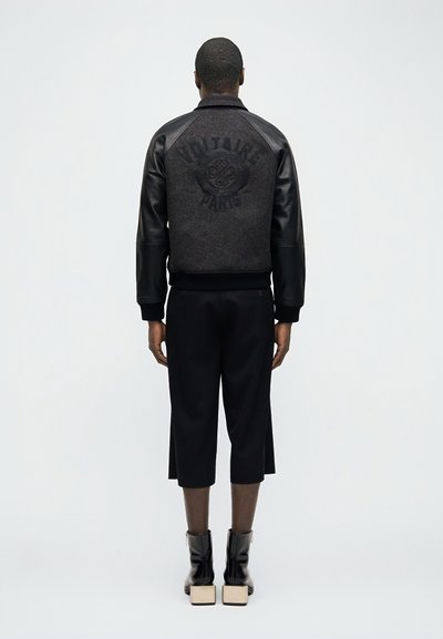 Άνδρας μοντέλο που φοράει ένα μαύρο και σκούρο γκρι μπουφάν Voltaire Paris, μαύρο παντελόνι μέχρι τον αστράγαλο και μαύρα μποτάκια με τετράγωνες μπεζ τακούνια, στέκεται γυρισμένος προς τα πίσω.