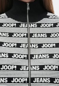 Black and white zip-up sweater with high collar, featuring repeated "JOOP!" and "JEANS" text. Knitted texture with striped pattern.