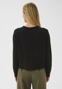 Frau mit langen blonden Haaren, die einen schwarzen Pullover und olivgrüne Hose trägt, von hinten vor einem einfarbigen weißen Hintergrund gesehen.