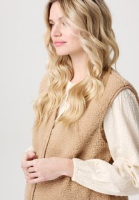 Beige fleeceväst med en mjuk textur, bärs över en vit, texturerad blus. Västen har en enkel design och rundad halsringning.