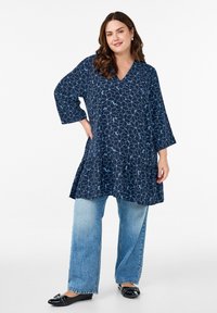 Navy blue tunic with white floral pattern, V-neckline, 3/4 sleeves, and ruffled hem, paired with light blue wide-leg jeans and black flats.