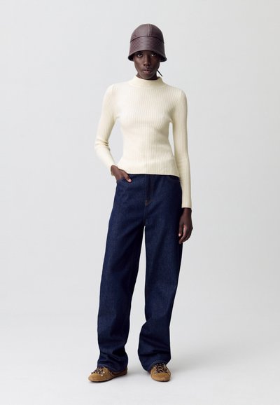 Modèle portant un haut à manches longues en série crème, un jean large foncé, des baskets beiges et un chapeau seau marron, se tenant devant un fond uni.
