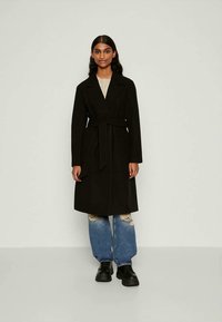 Casaco preto, longo, com um design envolvente e cinto, feito de tecido texturizado. Usado sobre uma blusa bege e combinado com jeans desgastados e sapatos pretos.