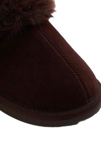 Zapatilla de ante marrón con punta redondeada, que cuenta con un acento de piel de pelo suave a lo largo del borde superior y una suela de goma oscura en contraste.
