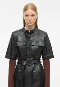 Juoda odinė trumpa suknelė trumpomis rankovėmis su priekinėmis kišenėmis, diržu ties juosmeniu ir apvaliu iškirpimu, derinama su bordo spalvos ilgarankove palaidine po ja.