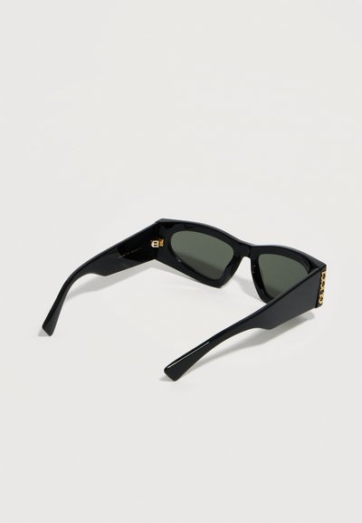 Μαύρα ορθογώνια γυαλιά ηλίου με σκούρους φακούς και λεπτομέρεια με χρυσό λογότυπο στα μπράτσα, ακουμπισμένα ανοιχτά πάνω σε ελαφρύ υπόστρωμα.