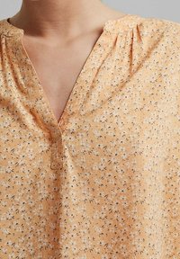 Ärmellose, orange Bluse mit V-Ausschnitt, mit einem floralen Muster in Weiß und Schwarz. Leichter Stoff mit feiner Textur.