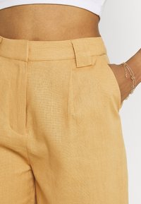 Light tan trousers made of textured fabric, featuring a flat front, side pockets, and belt loops. Tailored fit with subtle pleats.