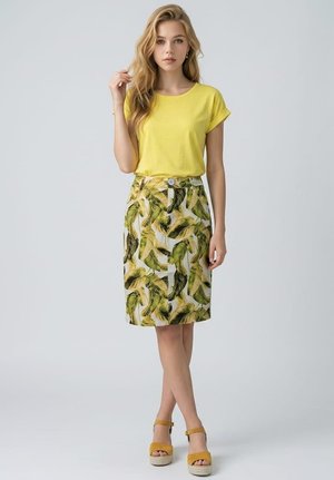 Joven mujer que lleva una camiseta amarilla, falda con estampado de hojas tropicales y sandalias amarillas con plataforma, de pie tocándose el cabello con una mano.