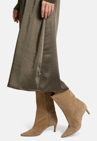 Unterbeine in taupe-farbenen Wildleder-Kitten-Heel-Stiefeln und einem langen olivgrünen Satinrock mit einem seitlichen Schlitz, die Hand ruht am Rock.