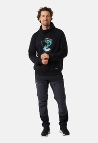 Haut à capuche noir avec impression graphique présentant un motif de poisson et de vague. Associé à un pantalon gris foncé avec poches zippées. Chaussures de sport décontractées.