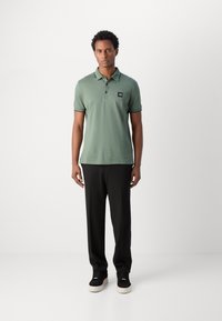 Groene polo met een korte kraag, zwart label op de linker borst, gecombineerd met zwarte broeken en zwarte sneakers, staande op een witte achtergrond.