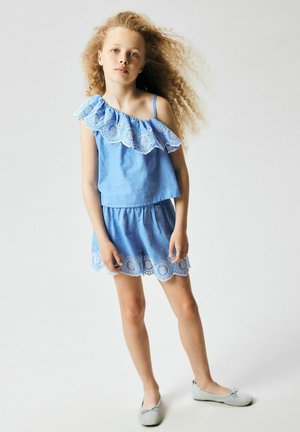 Joven niña con cabello rubio rizado, vistiendo un top azul sin hombros y una falda a juego con detalles de encaje blanco, posando frente a un fondo liso.