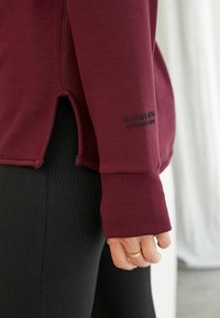 Bordeauxfarbenes Langarmoberteil mit einem Ausschnitt am Saum, ausgestattet mit gerippten Manschetten und einem schwarzen Textlogo "ELEMENTS COLLECTION" am Ärmel.