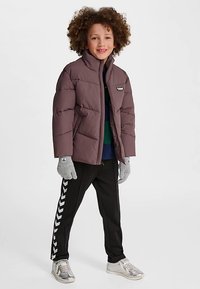 Doudoune violette foncée à coupe droite, gants gris, pantalons noirs ornés d'un motif en chevron blanc, et baskets argentées.