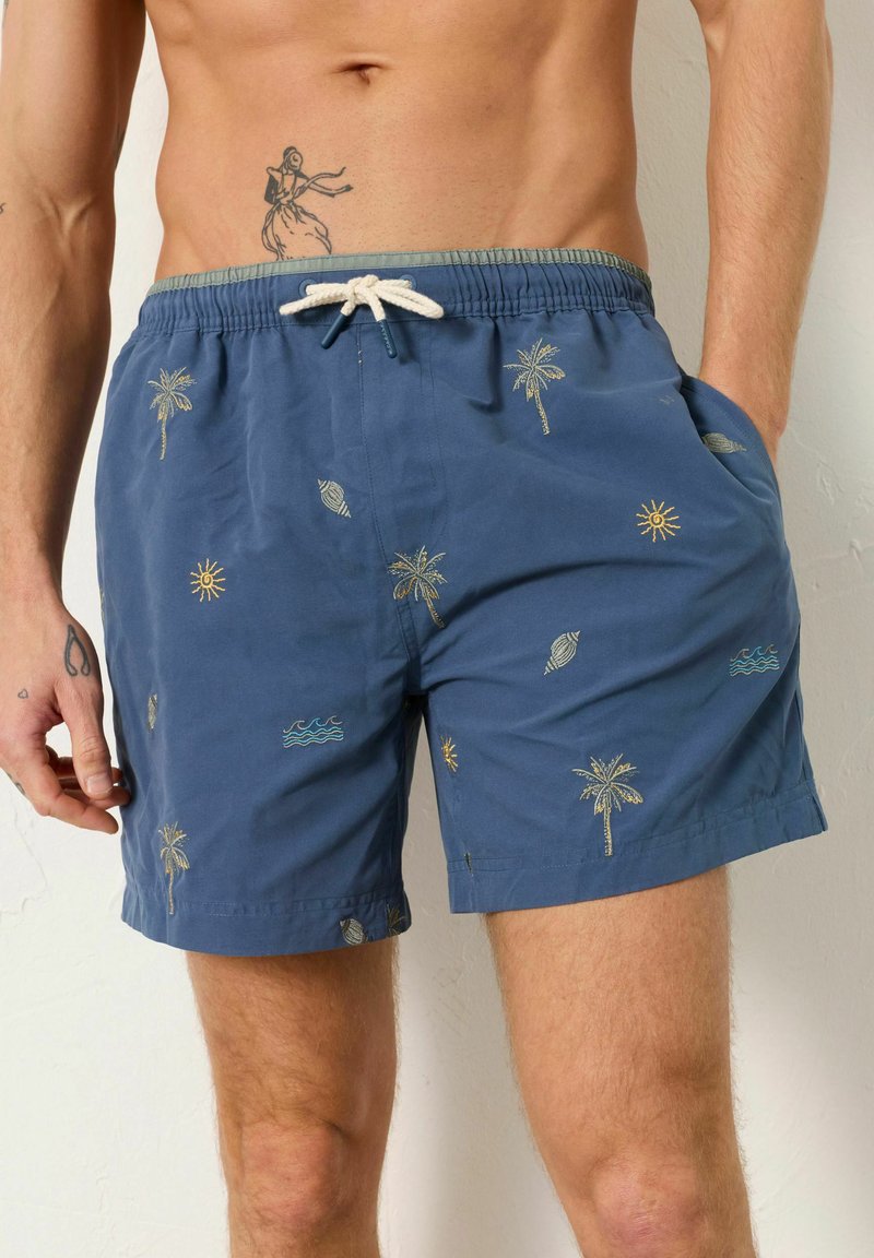 Blaue Badeshorts mit Mustern aus Palmen, Sonne, Muscheln und Wellen, getragen von einer Person mit Tätowierungen an Hand und Oberkörper.