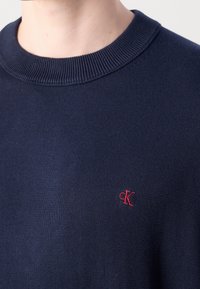 Gros plan d'une personne portant un pull bleu marine avec un logo brodé "cK" rouge près de la poitrine.