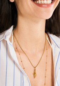 Collares en tono dorado con capas que presentan un charm de mano y acentos de cuentas delicadas, llevados sobre una camisa de rayas azules con un cuello clásico.