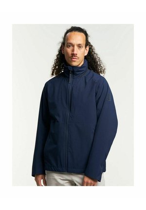 Man met krullend haar die een donkerblauw jasje met rits en lichte broek draagt, staand tegen een effen lichte achtergrond.