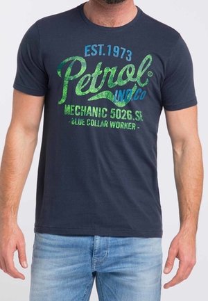 Homme portant un t-shirt bleu marine avec un texte vert et bleu indiquant "Petrol Inc Co Mechanic 5026 SI Blue Collar Worker" et un jean bleu clair.