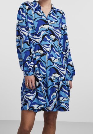 Robe bleue à manches longues avec motifs abstraits de vagues blanches et noires, devant boutonné, taille froncée et jupe à volants portée par une personne debout.