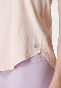 Ljust rosa, kortärmad topp av mjuk bomull med rundad kant och en liten logga på sidan; matchad med ljuslavendelfärgade leggings.
