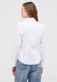 White button-up shirt with long sleeves, featuring a pointed collar, back yoke, and cuff buttons; paired with light blue denim jeans.