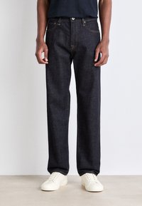 Pantalones de mezclilla azul oscuro con un diseño de pierna recta, que presentan costuras visibles, un cierre de botón y bolsillos frontales. Llevados con zapatillas blancas.