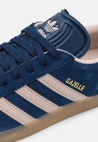 Blå mockasinsneaker med beigefärgade ränder, strukturerad yta och vadderad tungspets med en logotyp. Guldtexten "GAZELLE" på sidan.