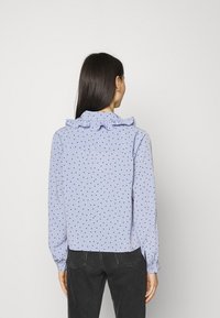 Blusa azzurro chiaro con pois neri, colletto arricciato, maniche lunghe e polsini con bottoni. Indossata con jeans scuri, vista da dietro.