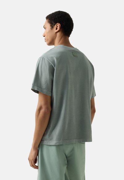 Mladý muž otočený doleva, oblečený v vybledlé zelené košili s krátkým rukávem a sladěných zelených kalhotách, na jednobarevném pozadí.