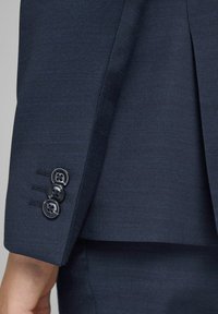 Une manche de veste de costume bleu marine avec un tissu texturé, trois boutons noirs, et une coupe ajustée avec de subtils détails de plis.