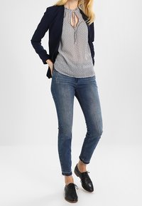 Blazer navy sopra una blusa bianca con un motivo, jeans skinny blu e scarpe nere con lacci. La blusa presenta un collo a chiave.