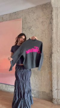 Femme debout à l'intérieur tenant un pull gris foncé avec un texte rose sur un cintre devant un mur texturé et un grand panneau rose.