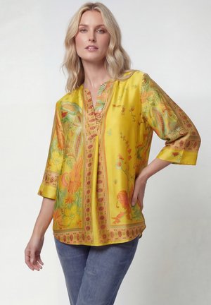 Blonde vrouw draagt een gele blouse met bloemenpatroon en driekwart mouwen en een blauwe spijkerbroek, poserend met één hand op haar heup.
