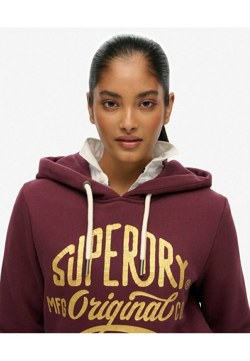 Bordowy hoodie z nadrukiem złotego logo, z kapturem na sznurki i białą koszulą pod spodem dla warstwowego stylu.