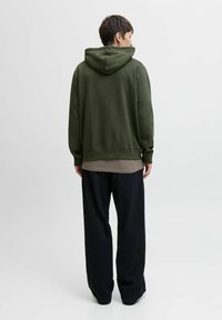 Dark green hoodie with a drawstring hood, relaxed fit, and ribbed cuffs, layered over a beige shirt, paired with wide black pants.