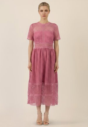 Woman standing wearing a pink short-sleeve midi dress with circular lace pattern and beige ankle-strap heels against plain background.