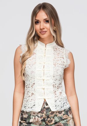 Jeune femme aux longs cheveux blonds porte un haut sans manches en dentelle blanche et un pantalon à motifs floraux sur fond gris uni.