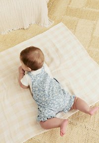 Blå blomsterprint overalls med bølgede kanter, båret over en hvid skjorte. Babyen ligger på et blødt, ternet tæppe på et struktureret gulv.