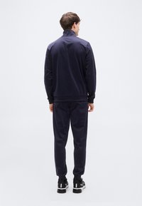 Σετ φόρμας σε ναυτικό μπλε με ζακέτα που κλείνει με φερμουάρ και ελαστικές μανσέτες, συνδυασμένο με παντελόνι στον ίδιο τόνο. Ομαλή υφή και μίνιμαλ σχεδιασμός. Μαύρα παπούτσια.