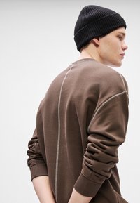 Brun sweatshirt med ribbstickade ärmar och rundaxel; har kontrasterande ljusa sömmar längs sömmarna. Bärs med en svart ribbstickad mössa.