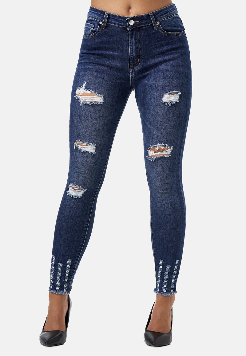 Jeans de mezclilla ajustados, de un azul oscuro, con detalles de rodillas rasgadas, dobladillos deshilachados y adornos en los tobillos. Textura suave, corte slim.
