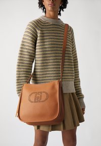 Borsa a spalla in pelle marrone con finitura texture, caratterizzata da un logo perforato. La tracolla è regolabile. Indossata con un maglione a righe.