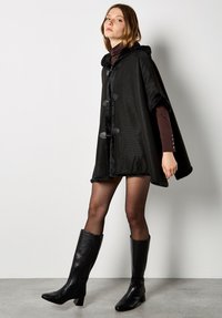 Schwarzer, strukturierter Cape mit Kapuze, ausgestattet mit Fellbesatz und Knebelverschlüssen. Wird über einem braunen Oberteil getragen und mit kniehohen schwarzen Stiefeln kombiniert.