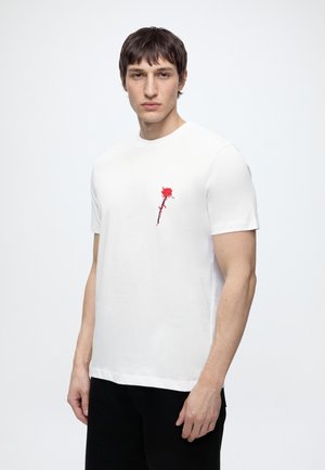 Mann trägt ein weißes T-Shirt mit kleinem rotem Rosendesign auf der linken Brust, steht vor einem einfarbigen hellen Hintergrund.