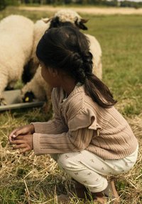 Pull en tricot rose clair avec un motif texturé, pantalon à motifs avec de petits nœuds, accroupie sur l'herbe près de moutons dans un cadre de ferme.